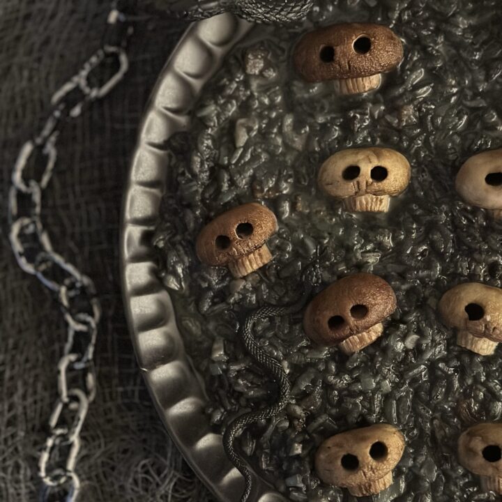 A shallow black serving plate filled with pitch black garlic mushroom risotto topped with creepy roasted button mushrooms carved into spooky skull shapes for a Halloween dinner party.