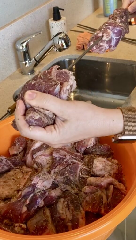 Global adventures in Darwin with friends teaching Chef Jen how to prep lamb for charcoals. a person is sliding large chunks of marinated lamb onto large metal skewers