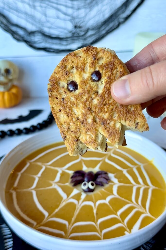 Chef Jen's hand holding a ghost grilled cheese over a bowl of butternut squash soup with a spiderweb pattern cream garnish and adorable olive spider resting on top with cute cartoon style eyes.