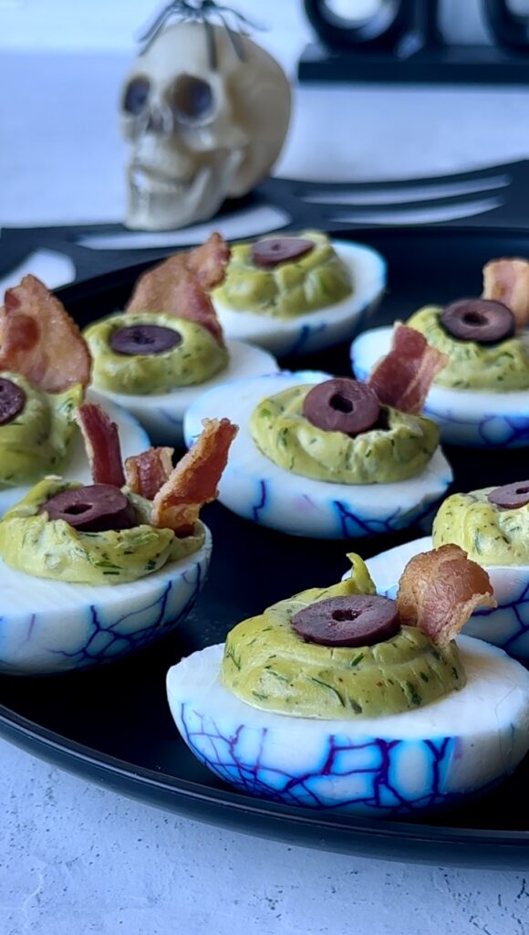 A black platter filled with completed viral zombie eyeball deviled eggs with purple veins, bright green filling as irises and bacon eyelids.