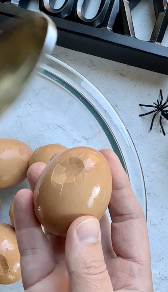 A close up shot of a chef's hand using the back of a spoon to gently tap fine, spiderweb-like hairline cracks into the shell of a brown hard-boiled egg over a clear Pyrex bowl.