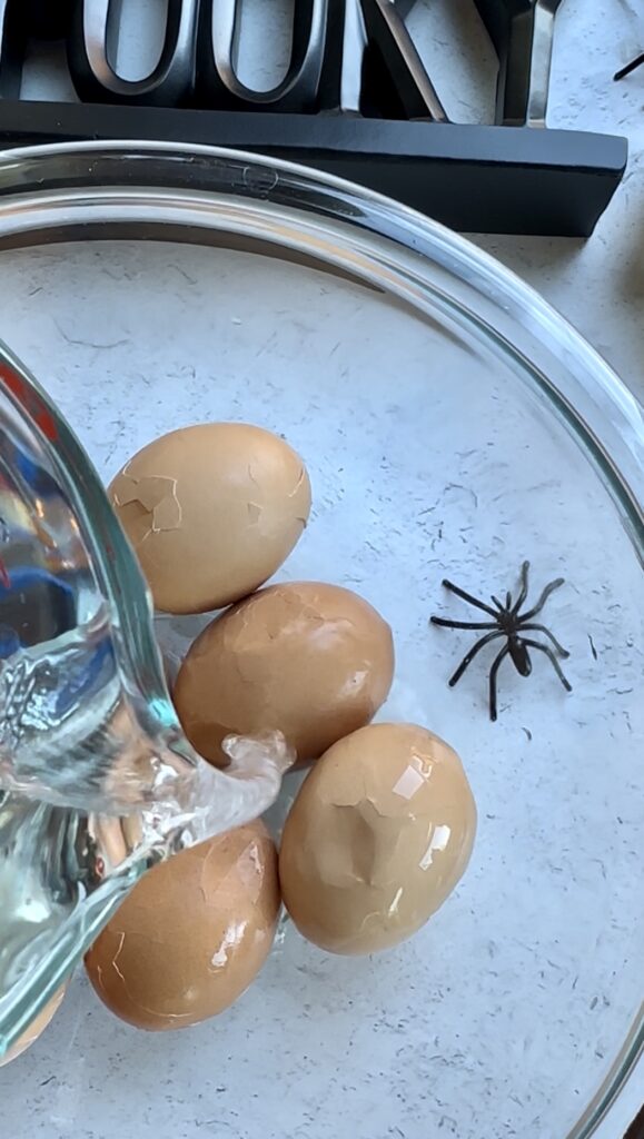 Cold water being poured into a Pyrex bowl to submerge cracked hard-boiled eggs, preparing the base for a spooky Halloween deviled egg appetizer with spiderweb pattern.