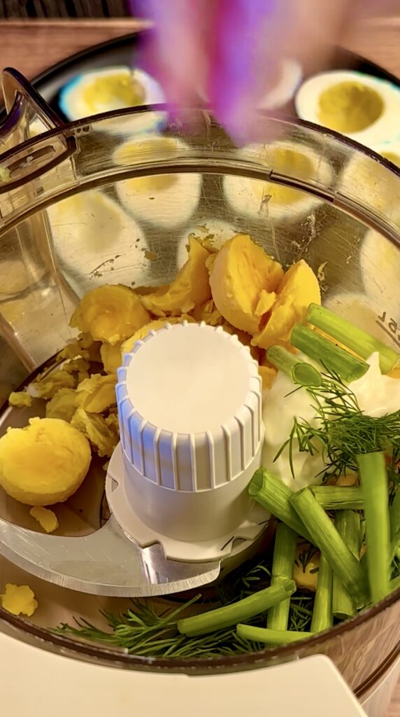 Hard-boiled egg yolks, fresh dill, green onion tops, and Dijon mustard inside a food processor bowl before blending.