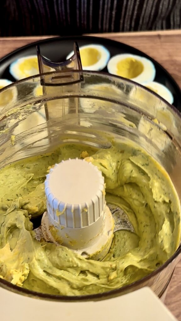 Creamy, smooth green goddess deviled egg filling blended inside a food processor bowl with fresh herbs.