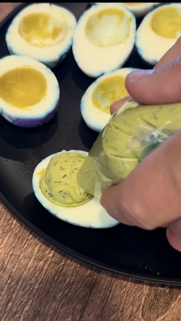 Piping a fresh green herbaceous filling into halved hard-boiled egg whites using a plastic Ziploc bag.