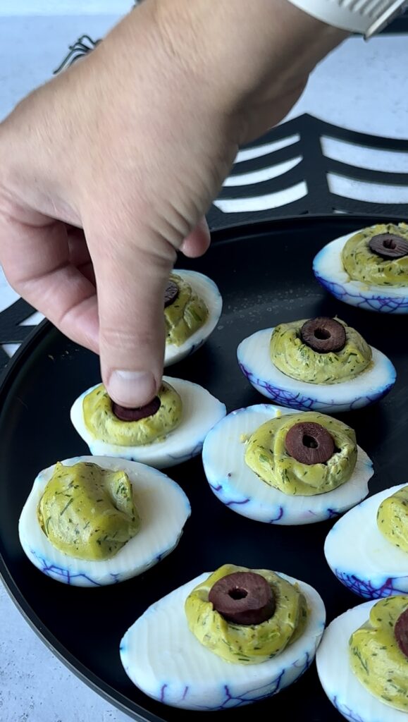 Placing a sliced black olive round as an eye pupil onto a green herbaceous Halloween deviled egg. 