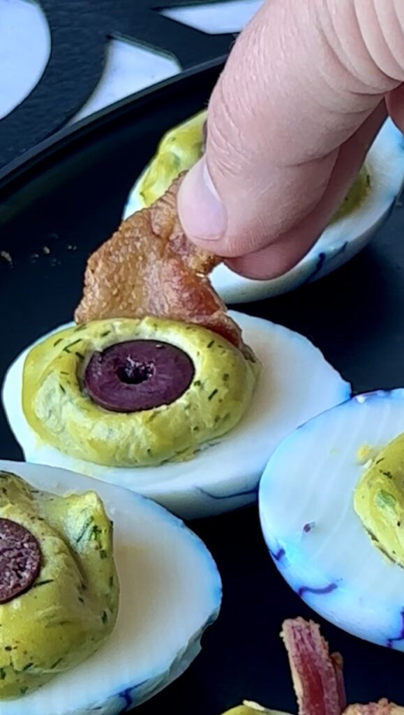 Adding a piece of crispy bacon over a black olive to create an eyelid or eyelash look on a green Halloween deviled egg.