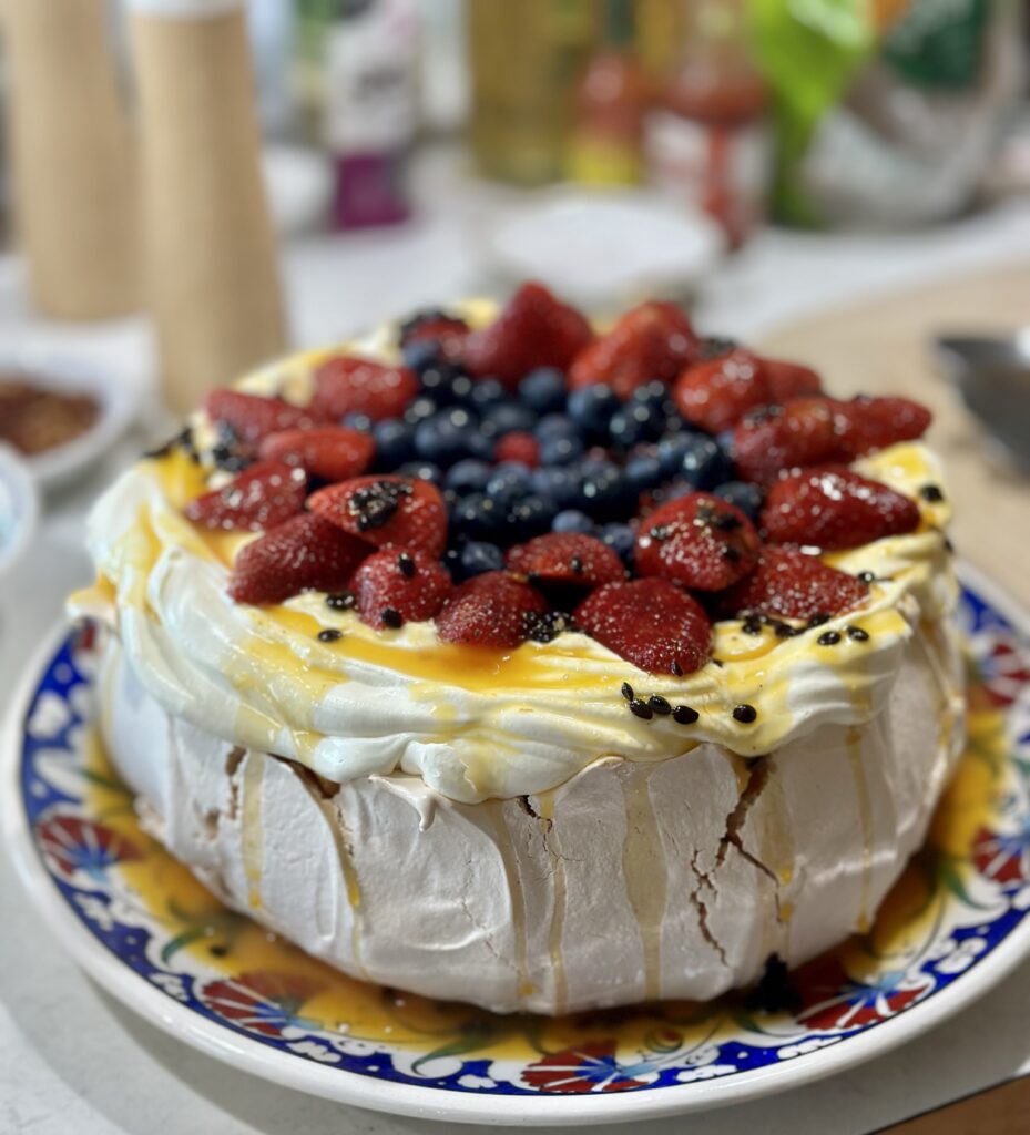 A beautiful pavlova slathered in whipped cream and covered in a generous arrangement of blueberries and strawberries with a passionfruit pulp drizzle on a colorful platter
