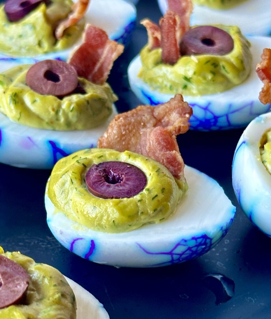The original viral zombie eyeball deviled eggs with a green goddess dill pickle filling, black olive slice pupil, and a crispy bacon shard garnish for a spooky Halloween appetizer.