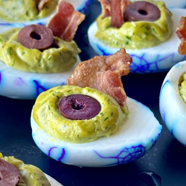 A matte black serving platter with spooky Halloween zombie eyeball deviled eggs featuring a vibrant purple spiderweb pattern on the egg whites and a bright green herb speckled filling appetizer