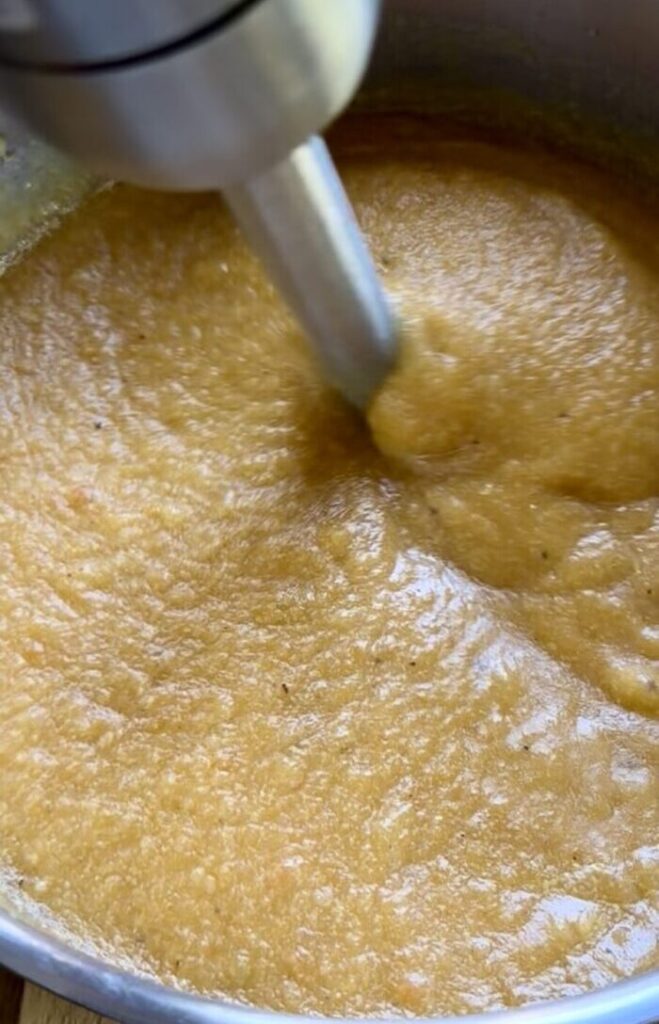 Blending cooked butternut squash and potatoes into a smooth velvety orange soup base using a hand immersion blender.