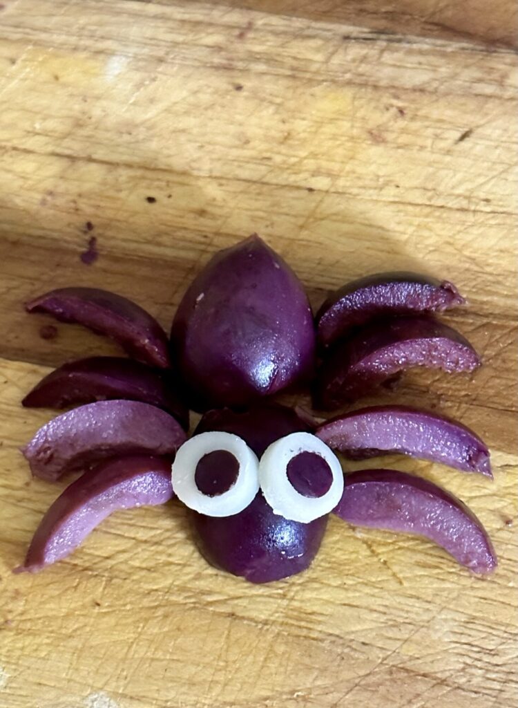 An adorable Halloween spider garnish made from black olives with provolone cheese eyes on a wooden board.