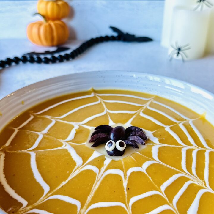 A perfectly centered overhead shot of a vibrant orange butternut squash spiderweb soup bowl with a cute decorative olive spider garnish floating in the center.