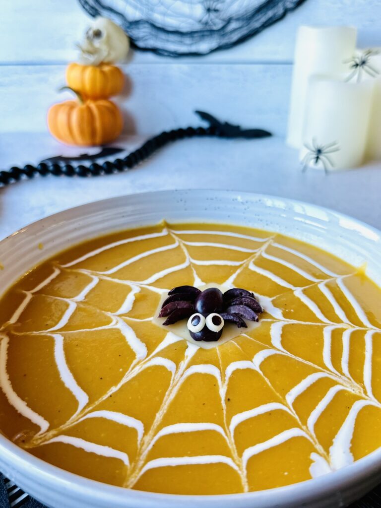 An adorable olive spider garnish floating perfectly on top of a provolone cheese island in the center of a cream spiderweb decorated orange soup. 