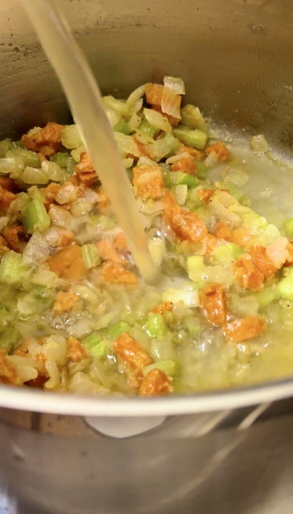 Pouring chicken stock into a pot with vegetables and scraping the bottom to deglaze