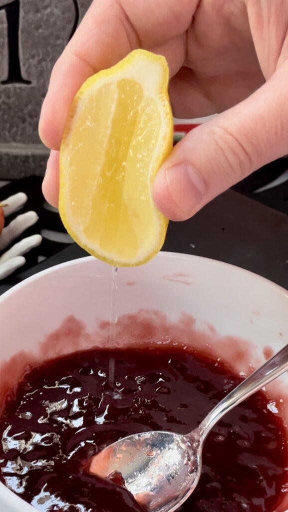 Adding fresh lemon juice to a homemade Halloween candy blood drip glaze. A hand squeezing a fresh lemon cheek into the red glaze to balance the sweetness.