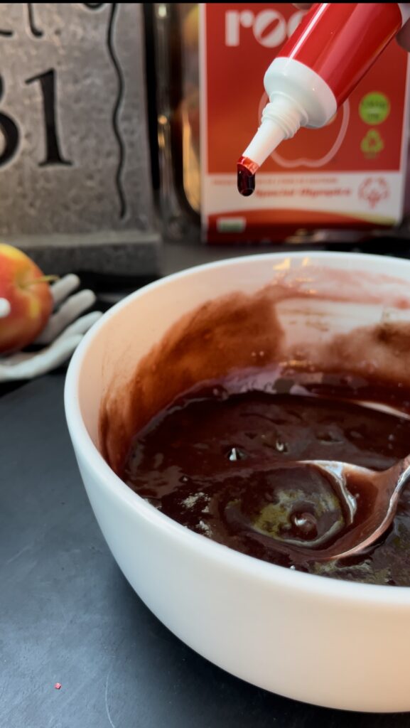 Deepening the color of the blood drip mixture with red food gel. A hand squeezing drops of vibrant red gel food coloring from a tube into a white mixing bowl of raspberry glaze.