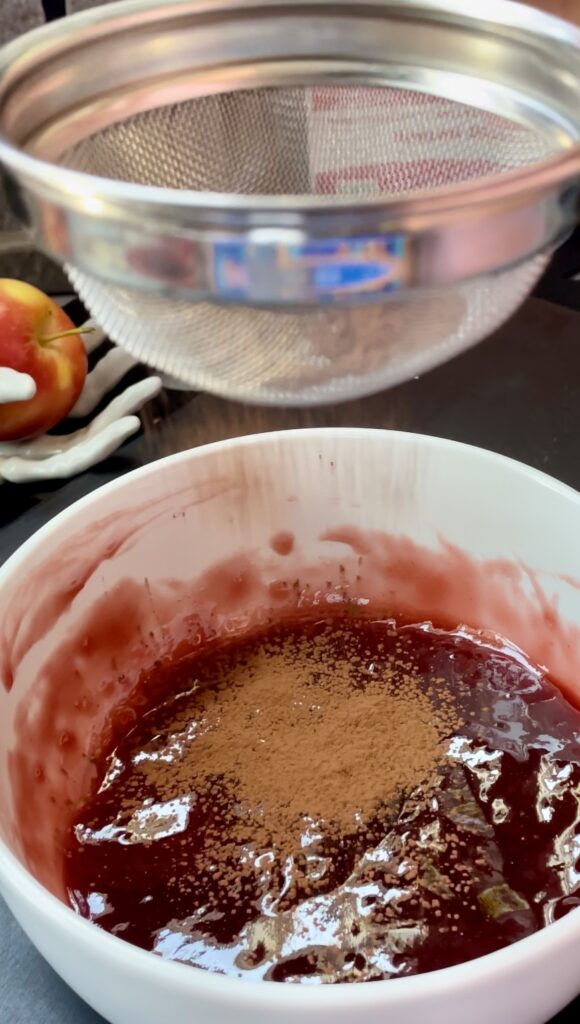 Dusting cocoa powder into a Halloween edible blood drip glaze to thicken and darken it. Sifting unsweetened cocoa powder through a metal mesh strainer into a bowl of glossy red raspberry glaze.