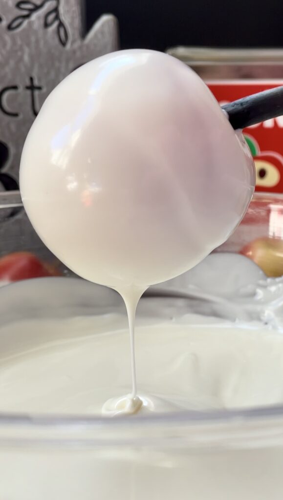 Smooth white chocolate dripping from a coated miniature apple into a mixing bowl. A close-up pf a miniature apple completely covered in smooth glossy white chocolate with a thick ribbon dripping down.