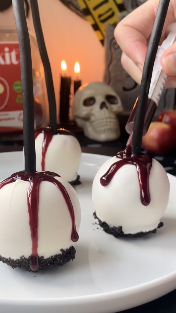 Applying realistic edible blood drips to Gothic Halloween candy apples with a syringe A hand using a plastic syringe to squeeze a thick glossy red blood glaze onto the top of white chocolate coated apples.