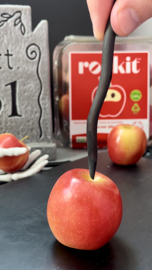 Inserting a black presentation stick at a straight angle into a miniature apple for a Halloween recipe. A close-up of a hand pressing a black stick straight down into the center stem cavity of a miniature apple on a clean black counter.