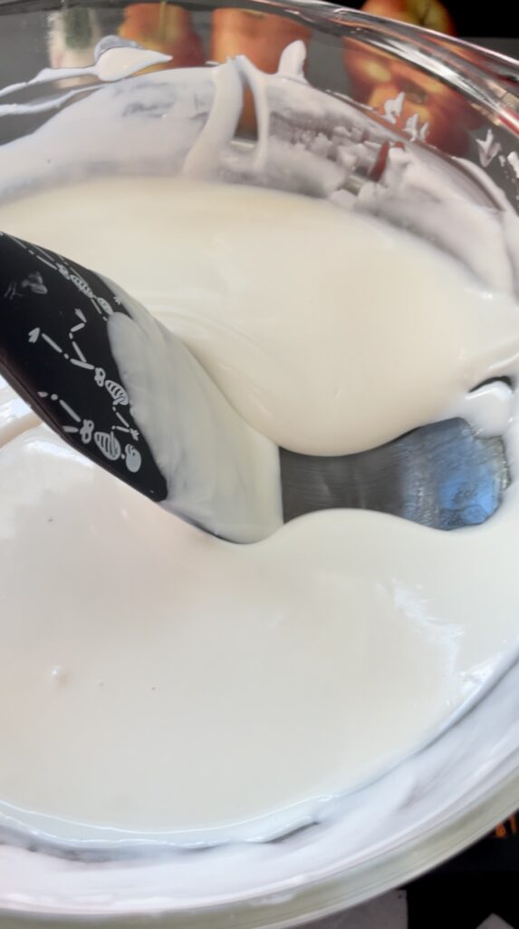 Checking the perfect coating thickness and ribbon viscosity of melted white chocolate. A black skeleton patterned spatula scraping and stirring melted white chocolate in a glass bowl to check the thickness.