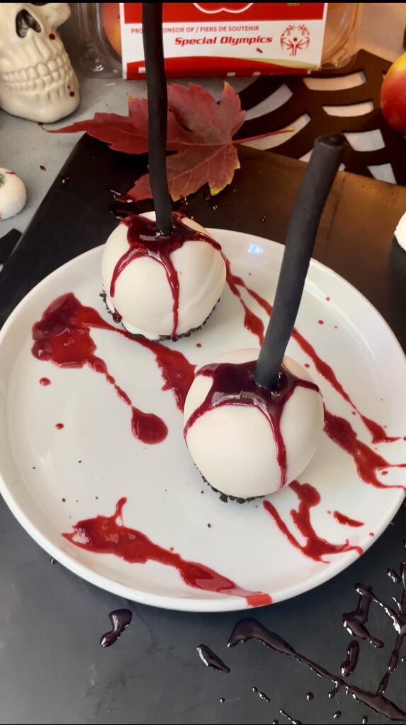 Spooky Halloween party table presentation ideas for bleeding white chocolate coated candy apples. A finished bleeding gothic white chocolate coated candy apple display showing two apples on black sticks with dripping blood and red splatter on a white plate.