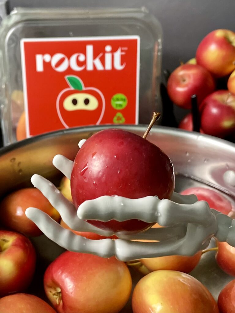Step-by-step tutorial for dewaxing mini apples for Halloween Gothic white chocolate candy apples. A plastic skeleton hand holding a miniature apple over a bowl of apples in water to clean off protective wax coating