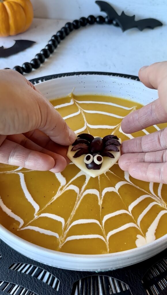 Hands gently lowering an olive spider garnish sitting on a round slice of provolone cheese buoy into a soup bowl.