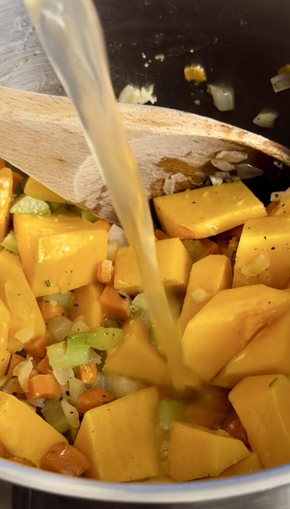 Pouring chicken stock over vegetables and fresh cubed butternut squash in a large cooking pot.